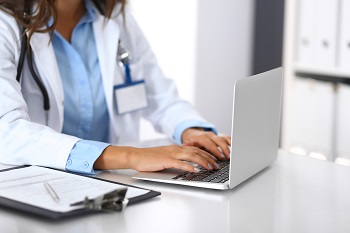 Female prescriber working on notebook computer with clipboard and pen by her side.